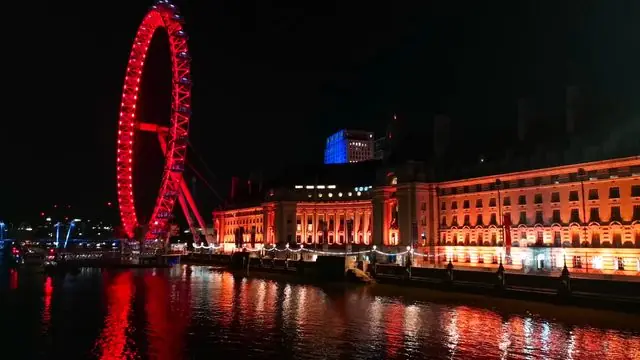 London Eye illuminated at night, reflecting in Thames with surrounding architecture.