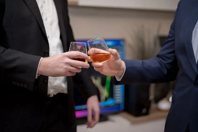 Two businessmen in suits toasting with drinks in an office environment.